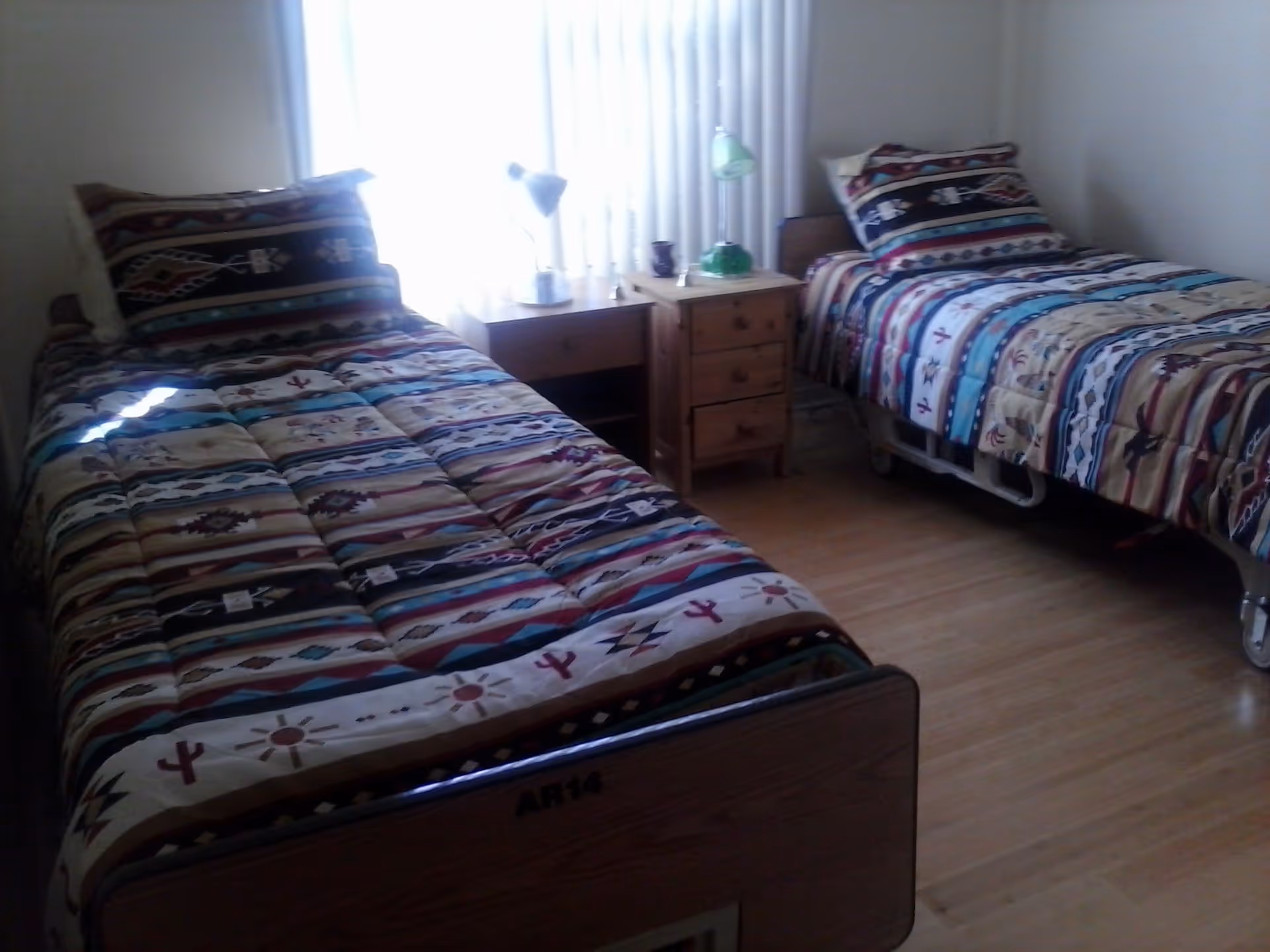 A simple bedroom with two single beds featuring patterned bedspreads separated by a small wooden nightstand beneath a window with vertical blinds.