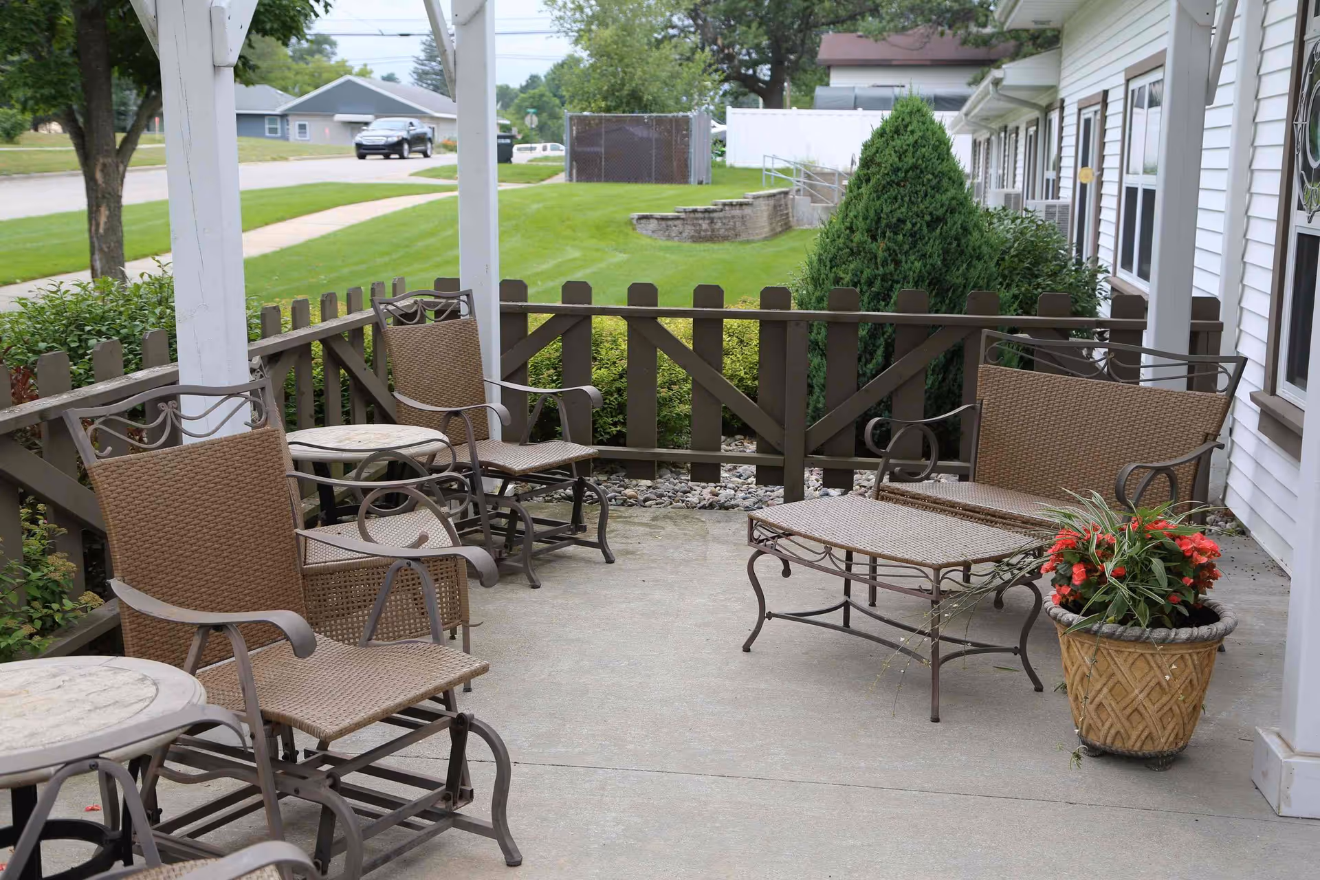 Outdoor patio seating area with wicker chairs, tables, a chaise lounge, and a potted flowering plant beside a fenced lawn.