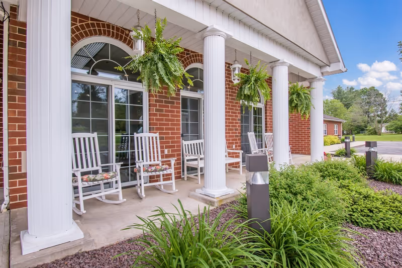 Front porch of a brick building with white columns, hanging green ferns, and white rocking chairs with floral cushions. There are glass doors behind the chairs and landscaped greenery in front of the porch under a blue sky with some clouds.