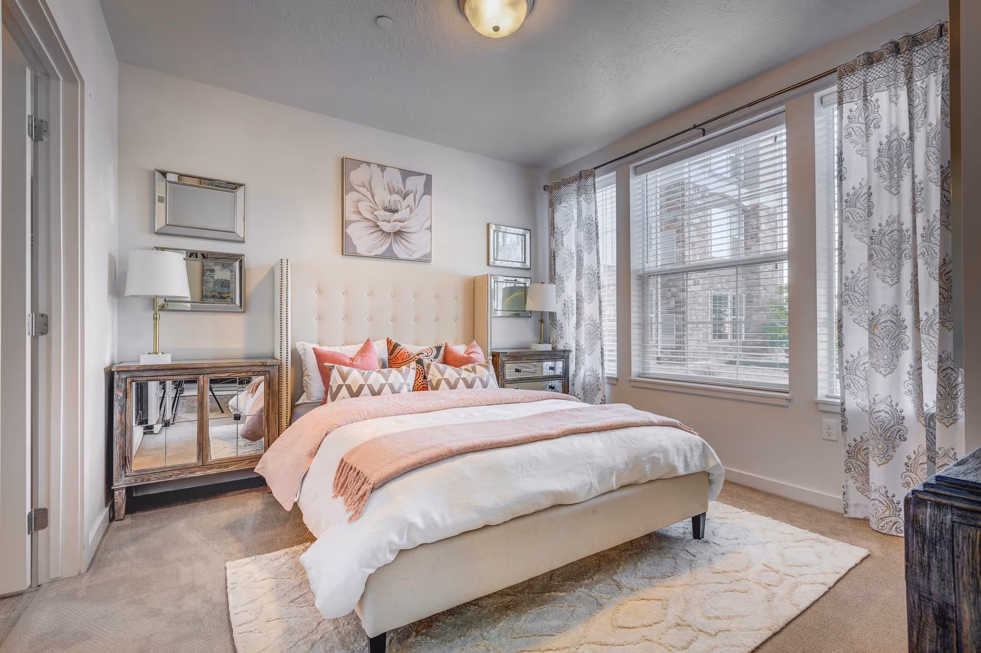 A bright and cozy bedroom with a large beige upholstered bed adorned with multiple decorative pillows and a pink throw blanket. The room features a soft beige carpet and a patterned area rug. On either side of the bed are mirrored nightstands with white lamps. The walls are decorated with framed artwork and mirrors. Large windows with white blinds and patterned curtains allow natural light to fill the room.