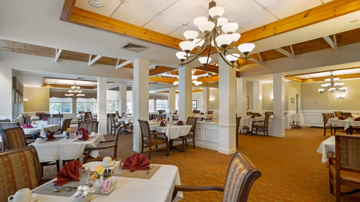 Spacious dining room with multiple tables covered in white tablecloths, each set with glassware, utensils, napkins folded in a decorative manner, and small flower arrangements. The room features carpeted floors, wooden ceiling beams, and multiple chandeliers providing warm lighting. Large windows allow natural light to enter the room.