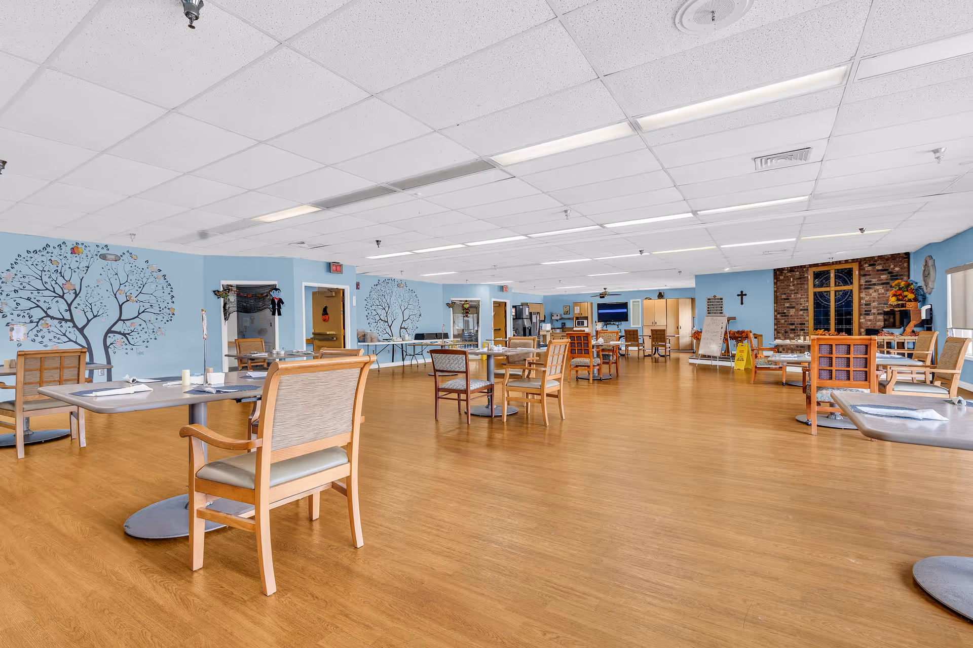 Spacious communal dining/activity room with scattered tables and chairs, blue walls featuring tree murals, and a wood floor.