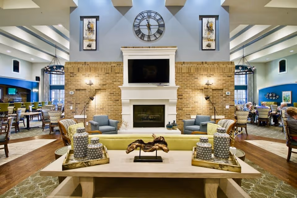 A spacious senior living facility common area with a central fireplace and mounted TV above it. The fireplace is flanked by two blue armchairs and wall lamps on a brick wall. In front of the fireplace is a wooden coffee table with decorative items and a green sofa. In the background, there are dining tables and chairs with seniors seated and socializing. The room has high ceilings with exposed beams and large windows allowing natural light.
