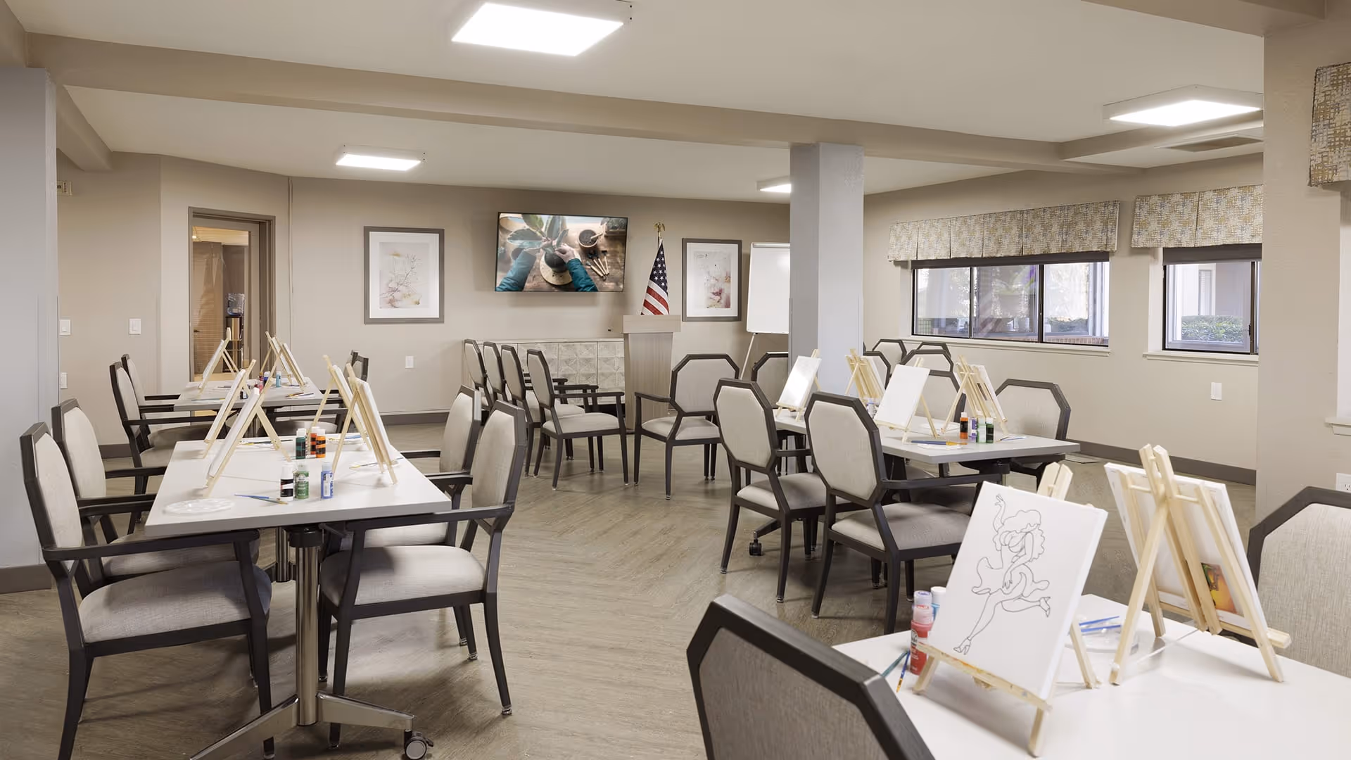 A well-lit activity room with several tables and chairs arranged for a painting session. Each table has small easels holding canvases, paint bottles, and brushes. The room has neutral-colored walls, large windows with valances, framed artwork, and an American flag near a podium. A TV is mounted on the wall displaying an image of paintbrushes and paint.
