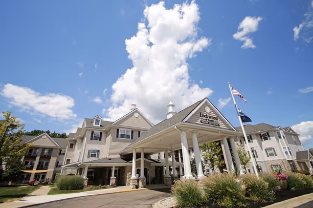 Exterior view of Lockwood of Fenton senior living facility under a blue sky with some clouds. The building has beige siding, multiple windows, and a covered entrance supported by white columns. There are American and other flags flying near the entrance, with landscaping including bushes and flowers around the driveway.