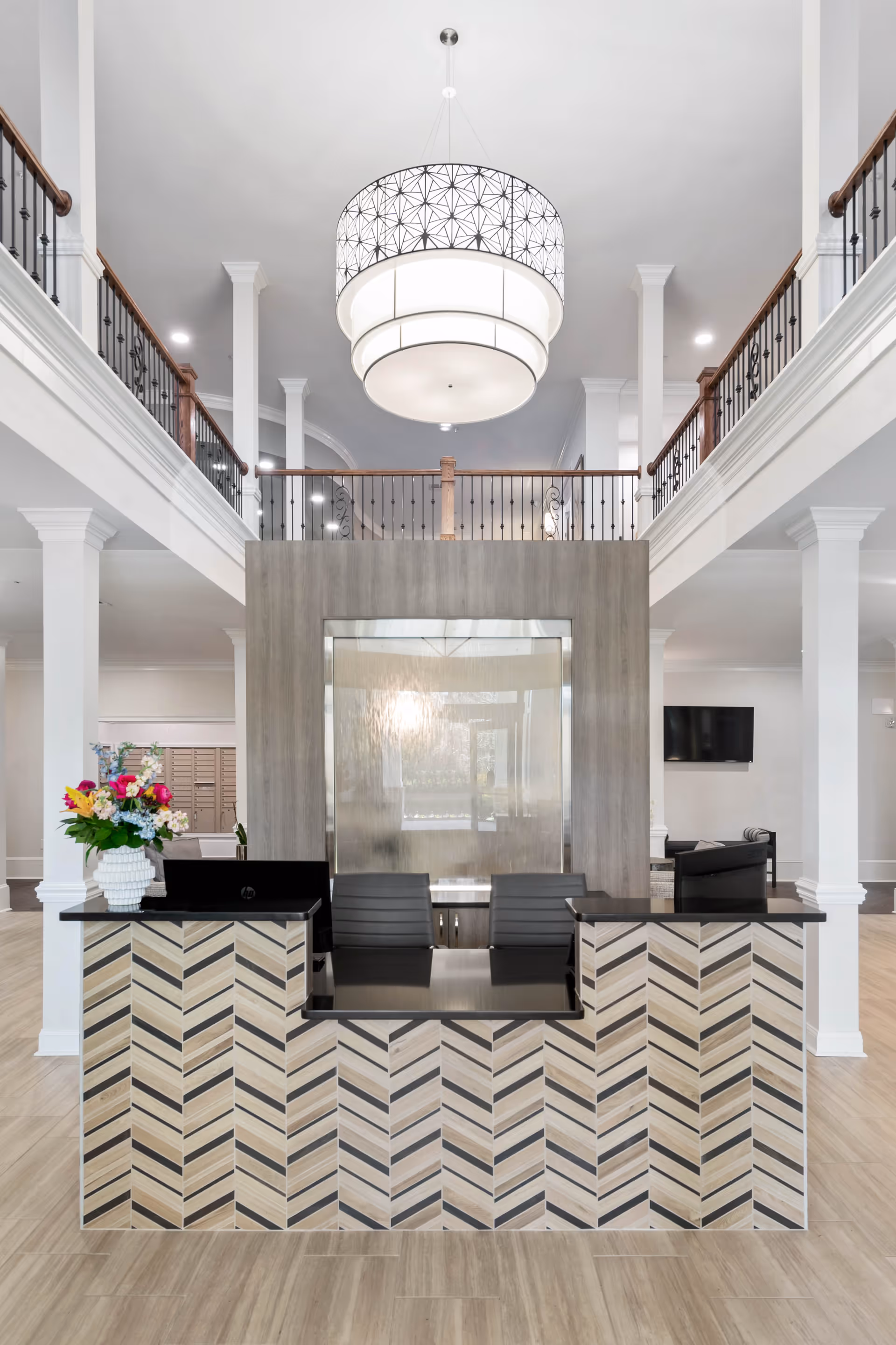 Reception area in a senior living facility with a modern front desk featuring a chevron pattern, two black chairs behind the desk, a large decorative hanging light fixture above, white columns, and a balcony railing on the second floor. A vase with colorful flowers is placed on the left side of the desk.
