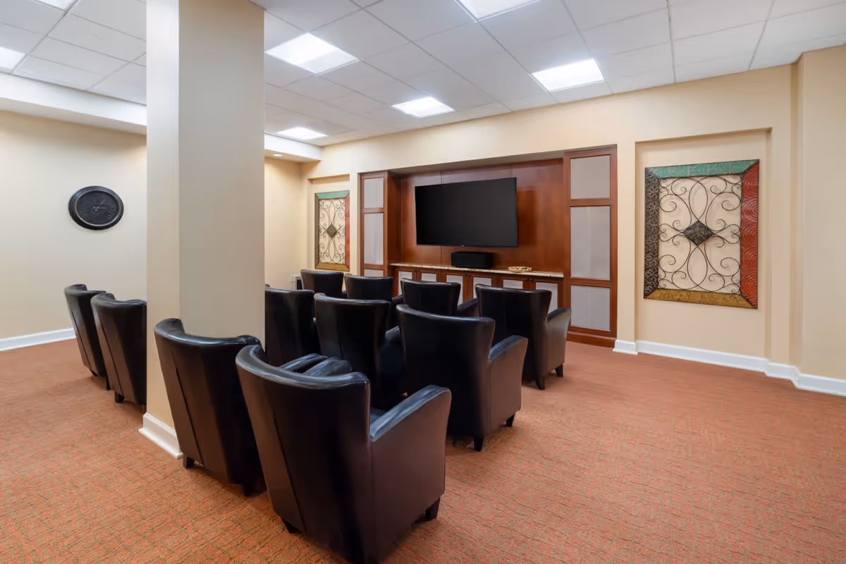 A small theater-style room with two rows of black leather armchairs facing a flat-screen TV mounted on a wooden entertainment center. The room has beige walls, a red carpet, decorative wall art on either side of the TV, and a clock on the left wall.