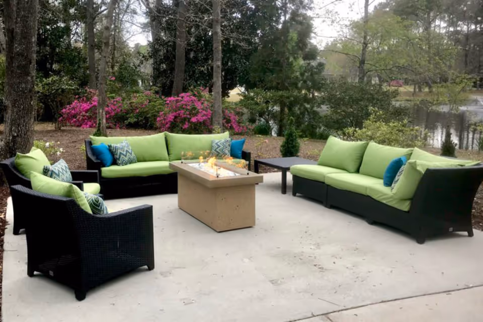 Outdoor seating area with black wicker furniture featuring green cushions and blue patterned pillows arranged around a rectangular fire pit table on a concrete patio. The background shows trees, pink flowering bushes, and a pond with a fountain.