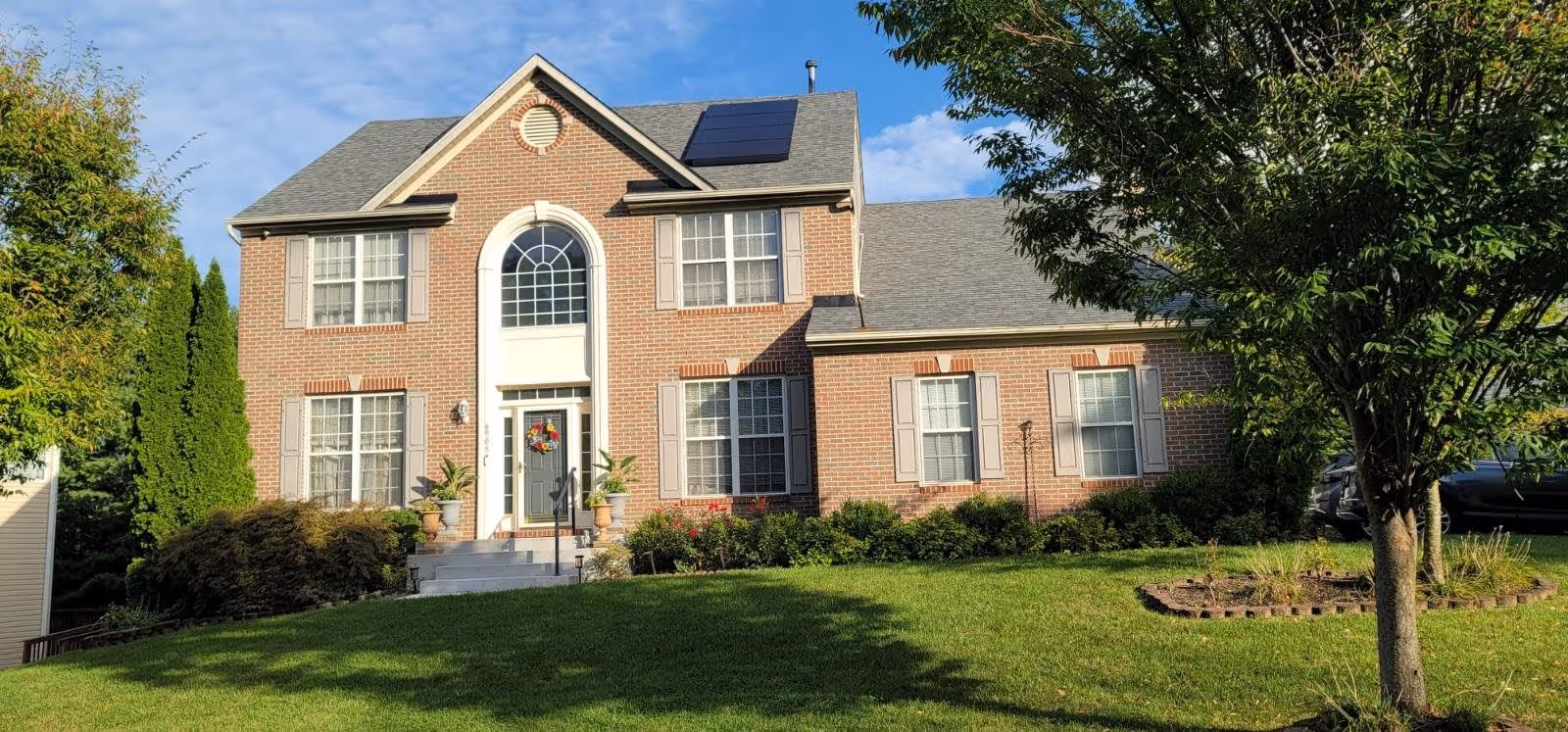 A two-story brick house with white trim and multiple windows, a front door with a decorative wreath, solar panels on the roof, surrounded by green grass, bushes, and trees under a partly cloudy sky.