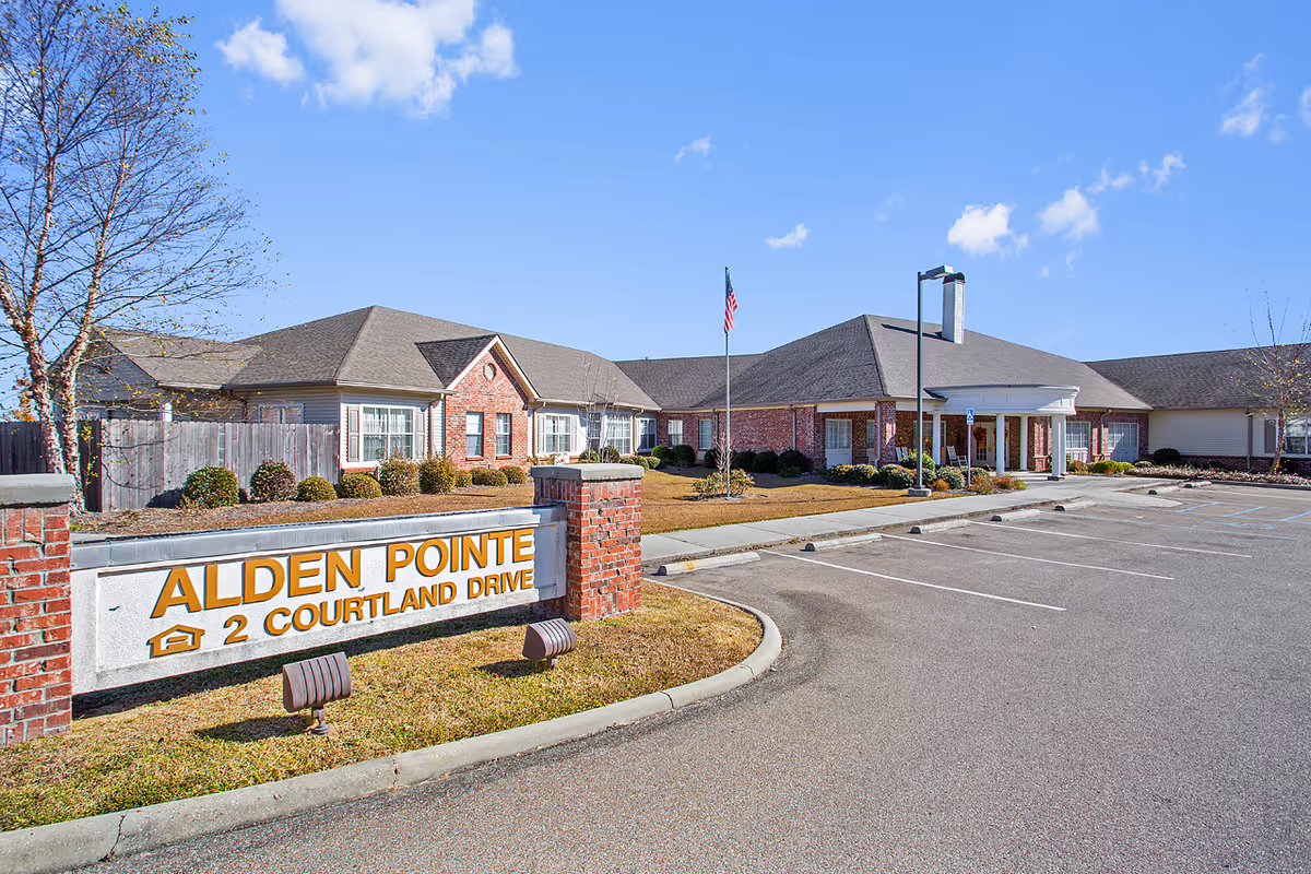 Exterior view of Alden Pointe Assisted Living & Memory Care facility showing a single-story brick building with a pitched roof, an American flag on a flagpole, a parking lot with marked spaces, and a sign in front that reads 'Alden Pointe 2 Courtland Drive'. The sky is clear with a few clouds.