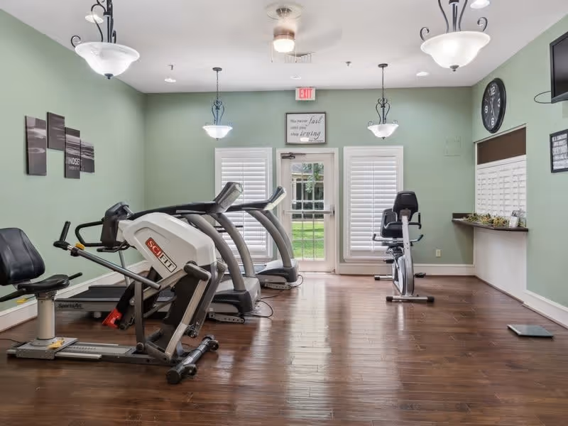 Small fitness room with treadmills, exercise bikes, and other cardio equipment facing a glass door and shuttered windows.