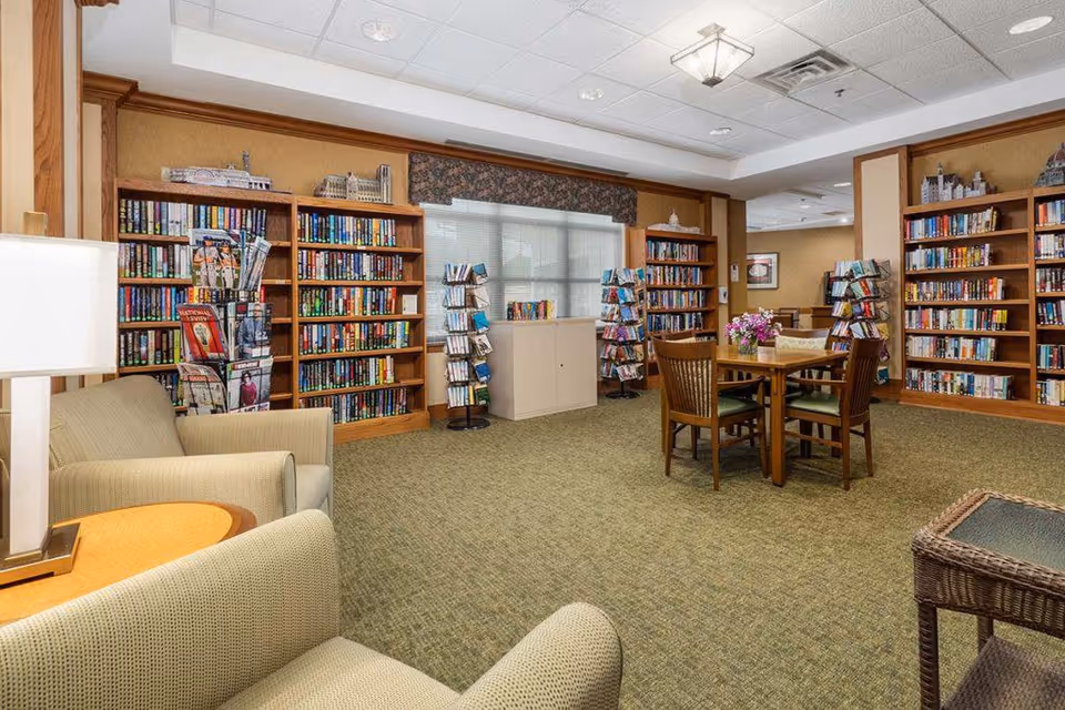 A cozy library or reading room with multiple wooden bookshelves filled with books and rotating racks of magazines or pamphlets. There are two beige armchairs with a small round wooden table and lamp between them in the foreground. In the center of the room, there is a wooden table with four chairs and a flower arrangement on top. The room has carpeted flooring and a window with blinds and a valance.
