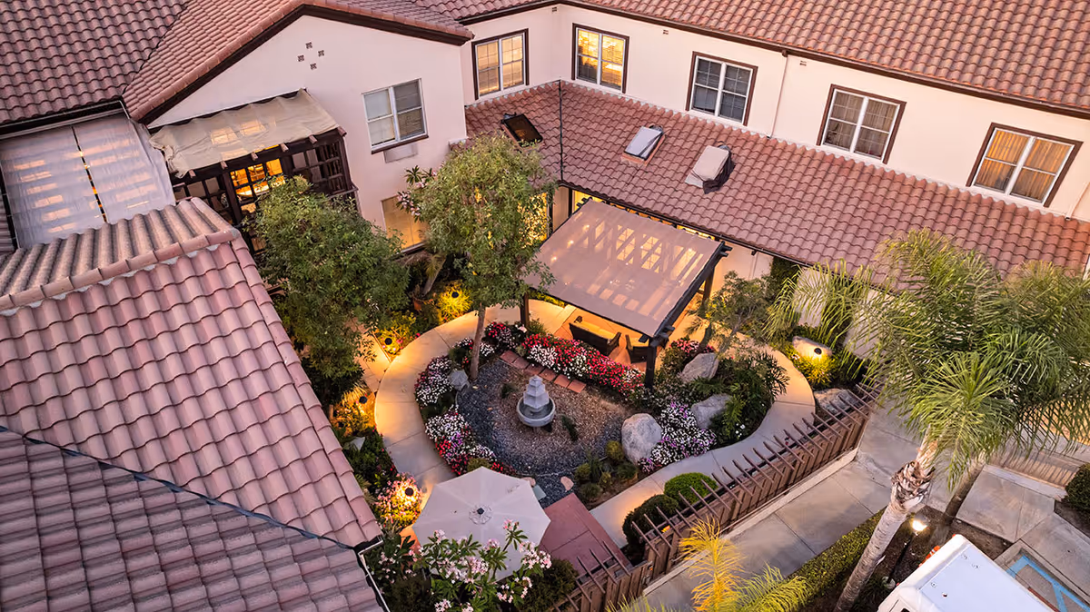 Aerial view of a courtyard garden at dusk within a senior living facility. The courtyard features a circular pathway surrounding a central fountain, colorful flower beds, trees, and a shaded seating area with a pergola. The surrounding building has multiple windows and a tiled roof, with outdoor lighting illuminating the garden.