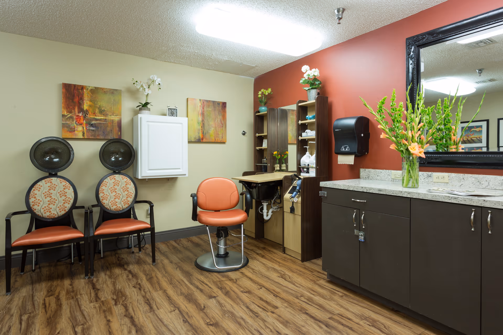 Interior salon area with two hooded hair dryers, an orange styling chair, countertop sink, cabinets and a large mirror.