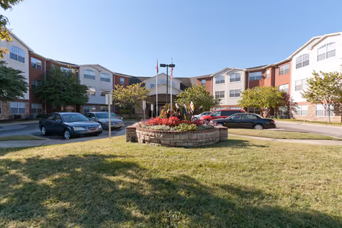 Exterior view of a senior living facility named Brookdale Franklin with a circular driveway, parked cars, a landscaped roundabout with flowers and trees, and a clear blue sky.