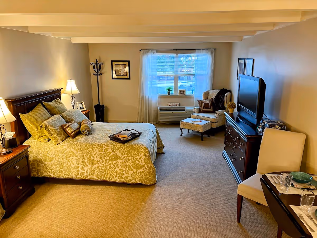 A cozy bedroom with a large bed covered in patterned bedding and multiple pillows. There are two bedside tables with lamps on each side of the bed. A window with sheer curtains lets in natural light. In front of the window is a cushioned armchair with an ottoman and a small side table. A dresser with a flat-screen TV on top is positioned against the wall, and a small dining table with two chairs is set with plates and glasses. The room has beige walls and carpeted flooring.