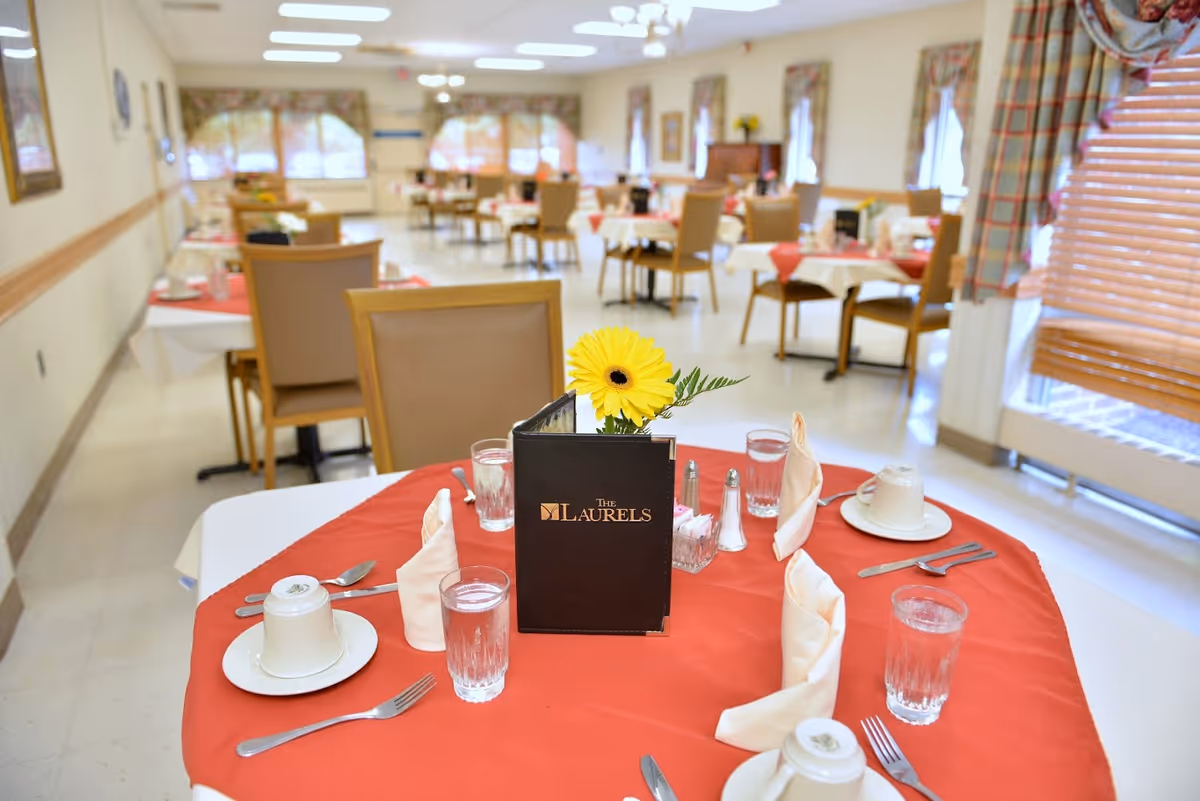 A dining room with tables set for a meal, featuring red tablecloths and white napkins folded upright. Each table has glasses of water, cups and saucers, silverware, and a small flower arrangement. The room has large windows with blinds and curtains, and several chairs are arranged around the tables.