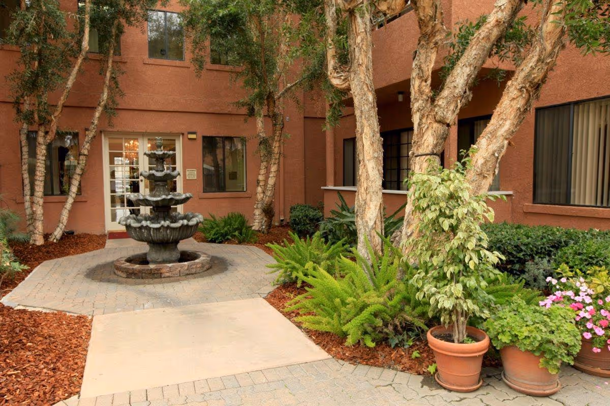 A landscaped courtyard with a three-tier fountain, potted plants and trees in front of a stucco building entrance.