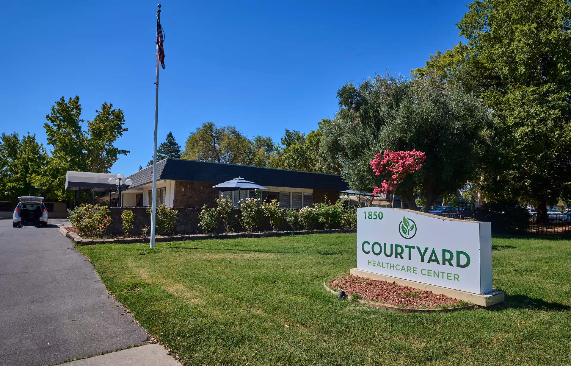 Exterior view of Courtyard Healthcare Center with a green lawn, a sign displaying the facility name and address, a flagpole with an American flag, and a building surrounded by trees and shrubs under a clear blue sky.