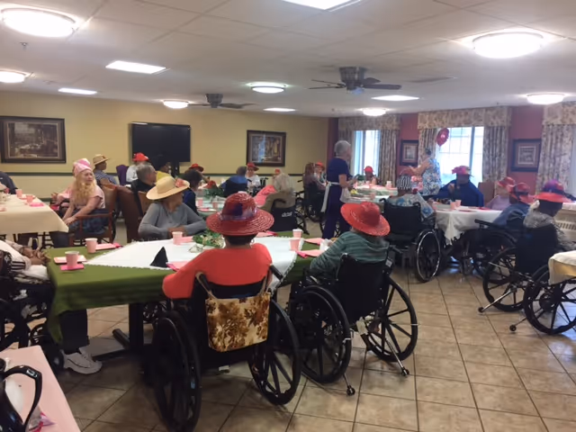 Several elderly residents in wheelchairs wearing red and pink hats seated around decorated tables in a community dining/activity room.