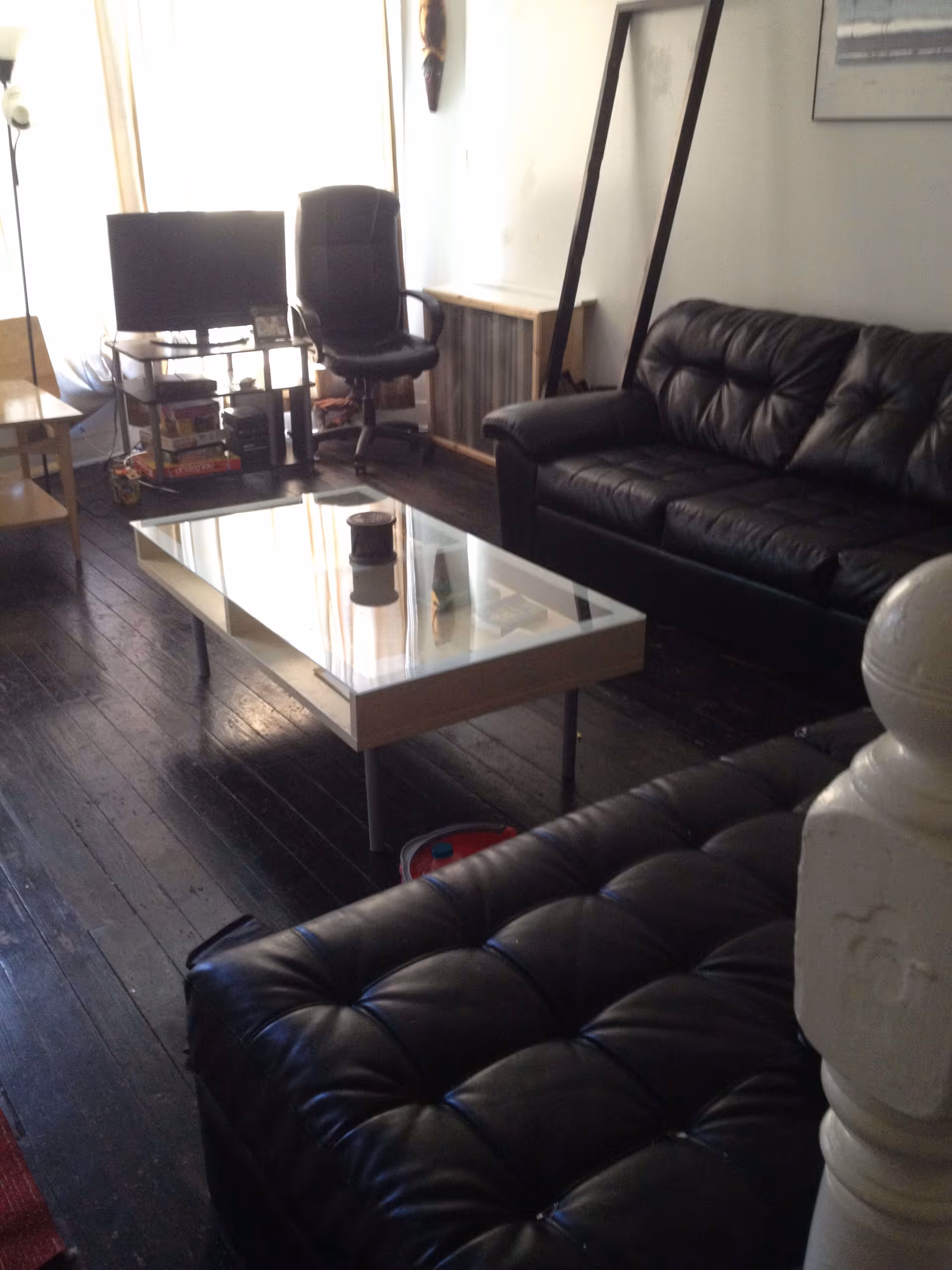Living room with black leather sofas, a glass-top coffee table, a TV on a stand and an office chair by a window.