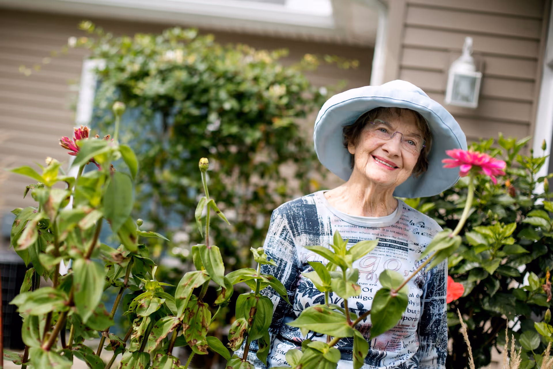 An elderly woman wearing a light blue wide-brimmed hat and glasses smiles while standing in a garden with green plants and pink flowers. The background shows part of a house exterior with beige siding and a white lantern light fixture.