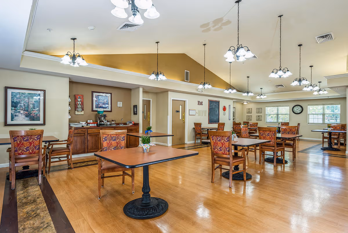A spacious dining room with multiple wooden tables and chairs featuring patterned upholstery. The room has a high ceiling with several hanging light fixtures, large windows letting in natural light, and framed artwork on the walls. There is a small counter area with coffee and other items in the back corner.