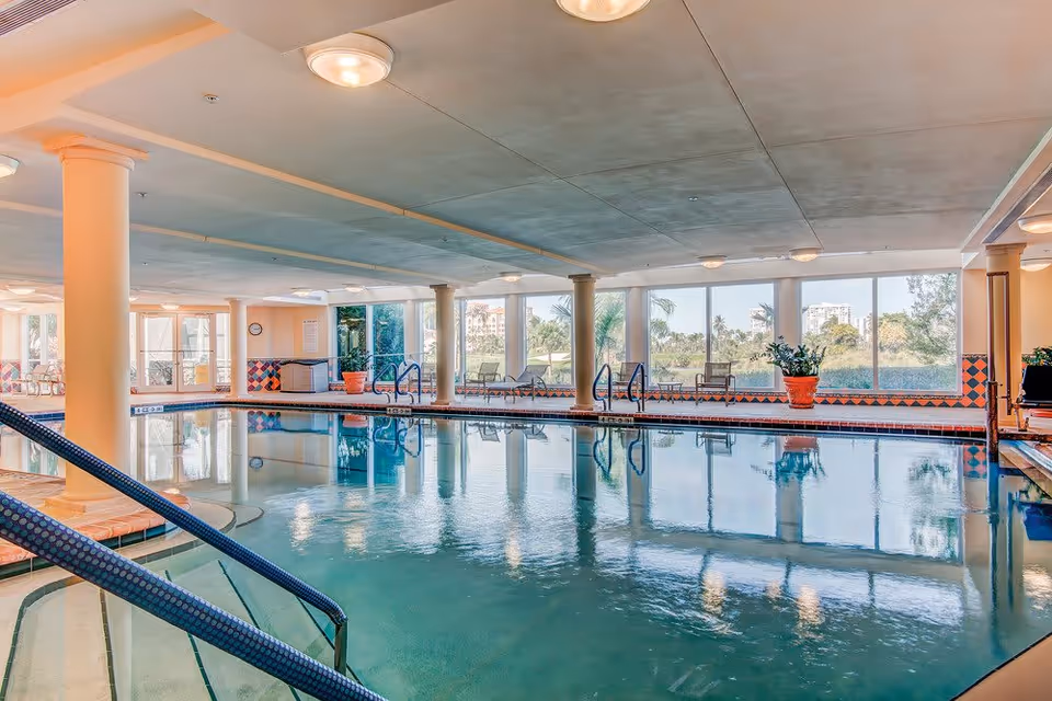 Indoor swimming pool area with large windows letting in natural light, several potted plants, pool handrails, and lounge chairs along the perimeter.