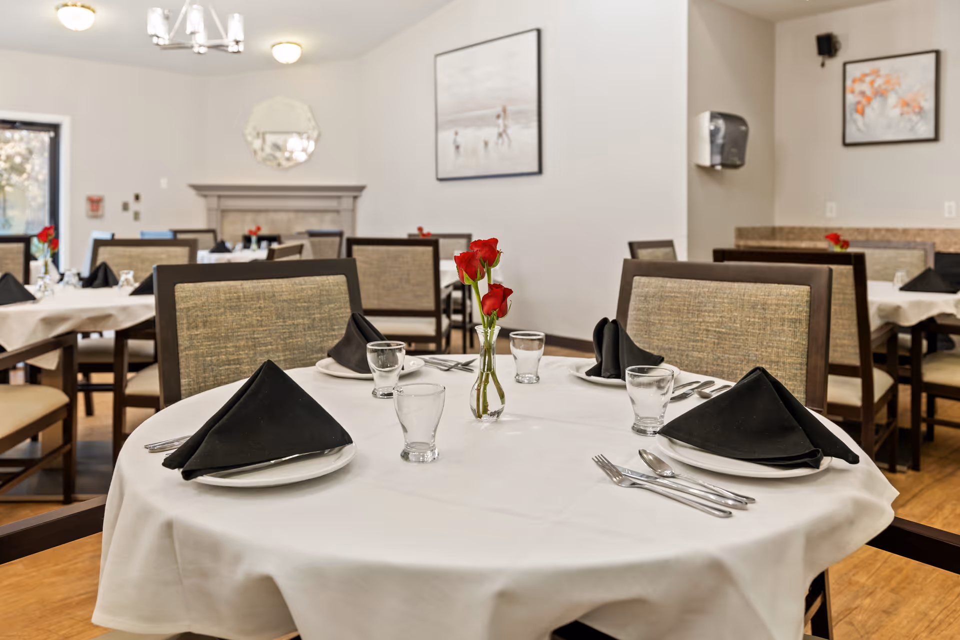 A dining room with a round table set with a white tablecloth, black napkins, glassware and a small vase of red roses, surrounded by chairs.