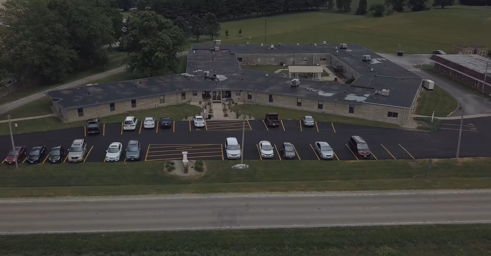 Aerial view of a single-story building with a flat roof, surrounded by a parking lot with several parked cars. The building is situated near a road and is surrounded by grassy areas and trees in the background.
