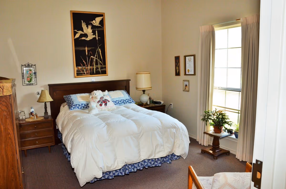 A cozy bedroom with a neatly made bed featuring a white comforter and decorative pillows, including a teddy bear pillow. The room has a wooden headboard, two bedside tables with lamps, framed artwork on the walls, a window with curtains, and a small table with potted plants near the window.