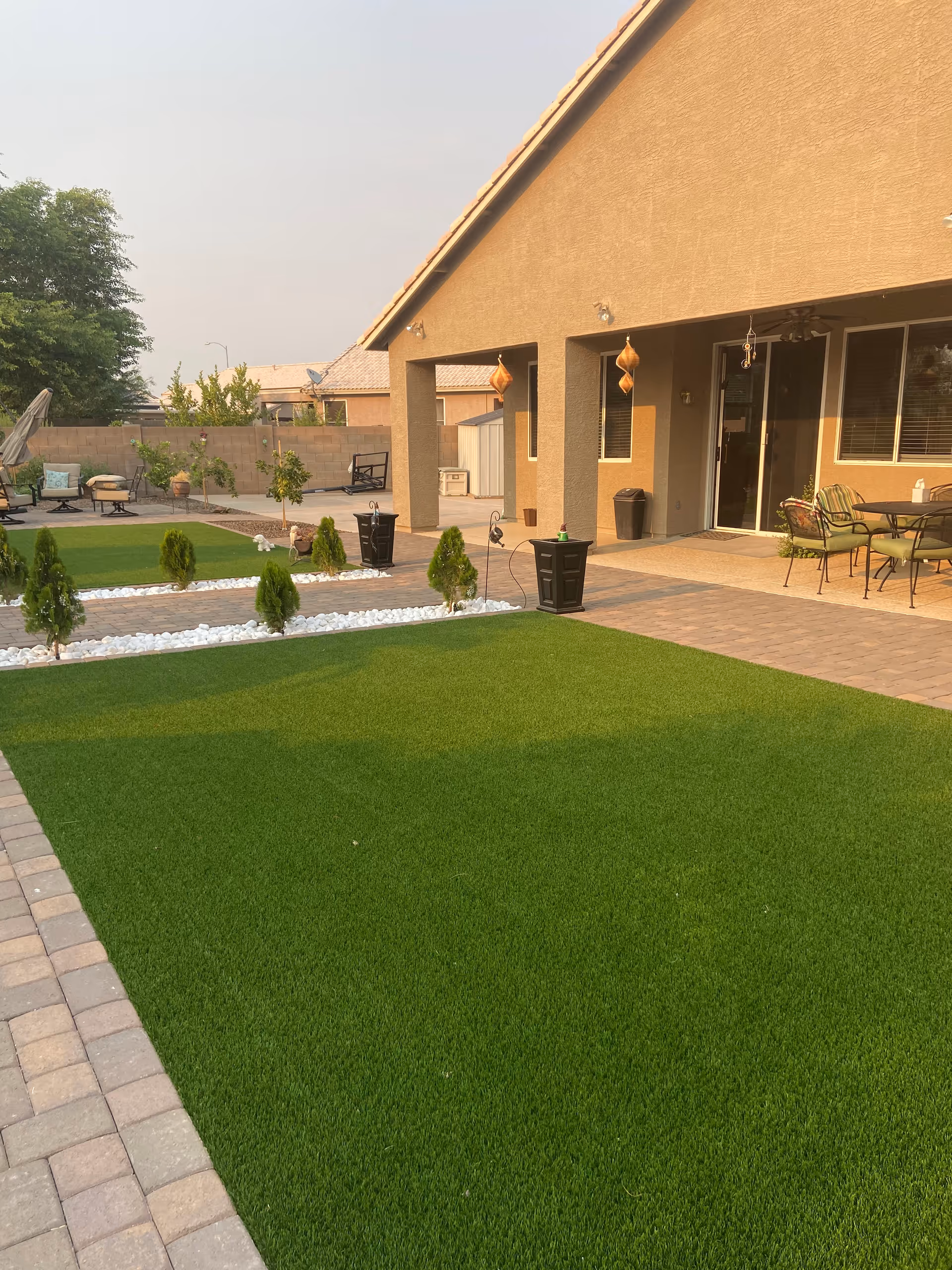 A backyard area with artificial green grass, small trees planted in white gravel beds, and a paved patio with outdoor furniture including a table and chairs. The house has a covered patio with hanging decorations and sliding glass doors.