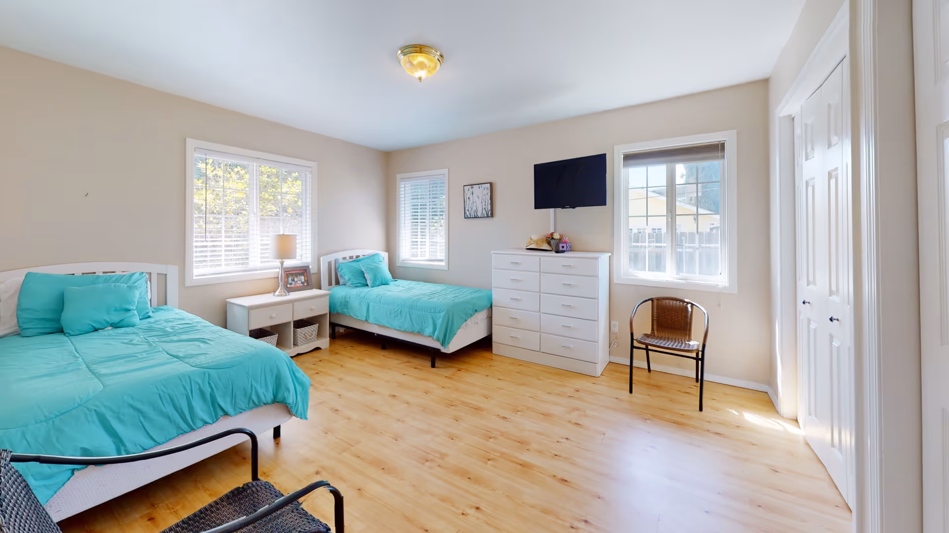 A bright bedroom with two single beds dressed in turquoise bedding, a white nightstand with a lamp and picture frame between the beds, a white dresser with a TV mounted above it, a wicker chair near the window, and light wood flooring. The room has beige walls and three windows letting in natural light.