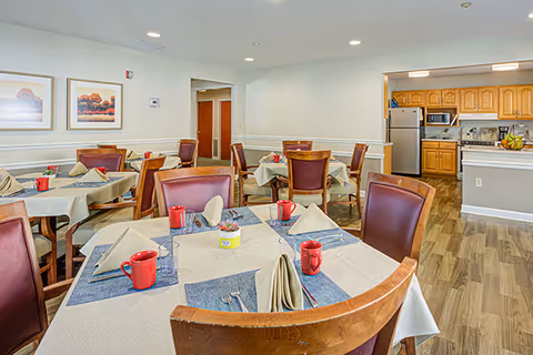 A dining room with several round tables covered with beige tablecloths and blue placemats. Each table is set with red mugs, glasses, silverware, and folded beige napkins. The room has wooden chairs with brown leather cushions. In the background, there is an open kitchen area with wooden cabinets, a refrigerator, microwave, and stove. The walls are light-colored with framed artwork and recessed lighting on the ceiling.