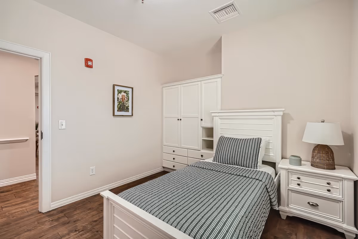 A simple, clean bedroom with a single white bed featuring a striped gray and white bedspread and pillow. Next to the bed is a white nightstand with a wicker lamp and a small decorative item. Behind the bed is a white wardrobe with drawers and shelves. The room has light-colored walls, wooden flooring, and a framed picture hanging on the wall. An open doorway leads to another room with similar flooring.