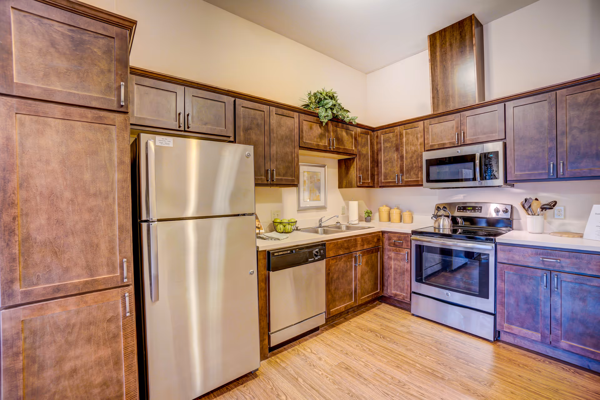 Modern kitchen with stainless steel refrigerator, dishwasher, oven, and microwave. The kitchen features dark wood cabinets, a double sink, and light-colored countertops. There is a small plant and decorative items on the counter, and the floor is wood.