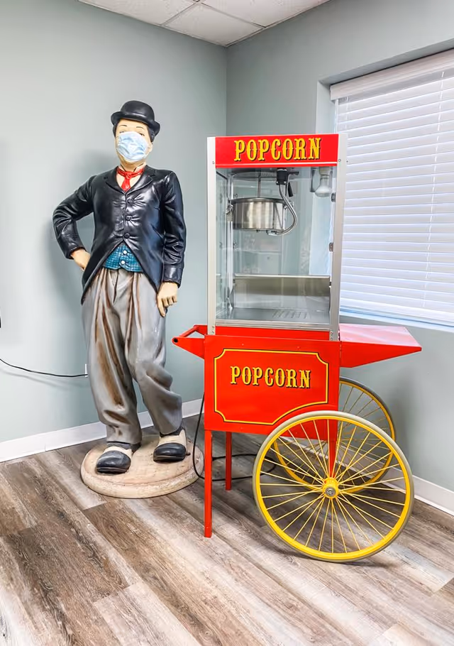 A life-sized statue of a man wearing a black bowler hat, black jacket, blue vest, gray pants, and black shoes, with a face mask on, standing next to a red popcorn machine with yellow wheels inside a room with light gray walls and wood-patterned flooring.