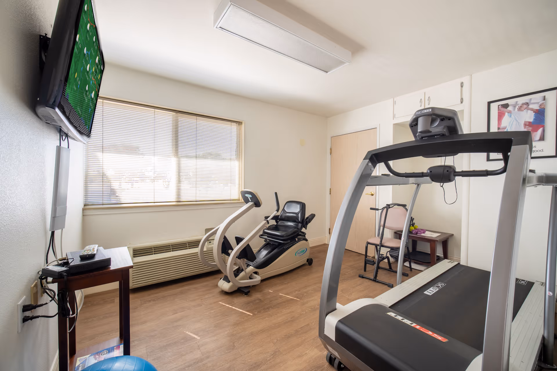 A small exercise room with a treadmill, a recumbent exercise bike, a chair, a small table with a plant, a wall-mounted TV, and a large window with blinds. The room has wooden flooring and white walls.