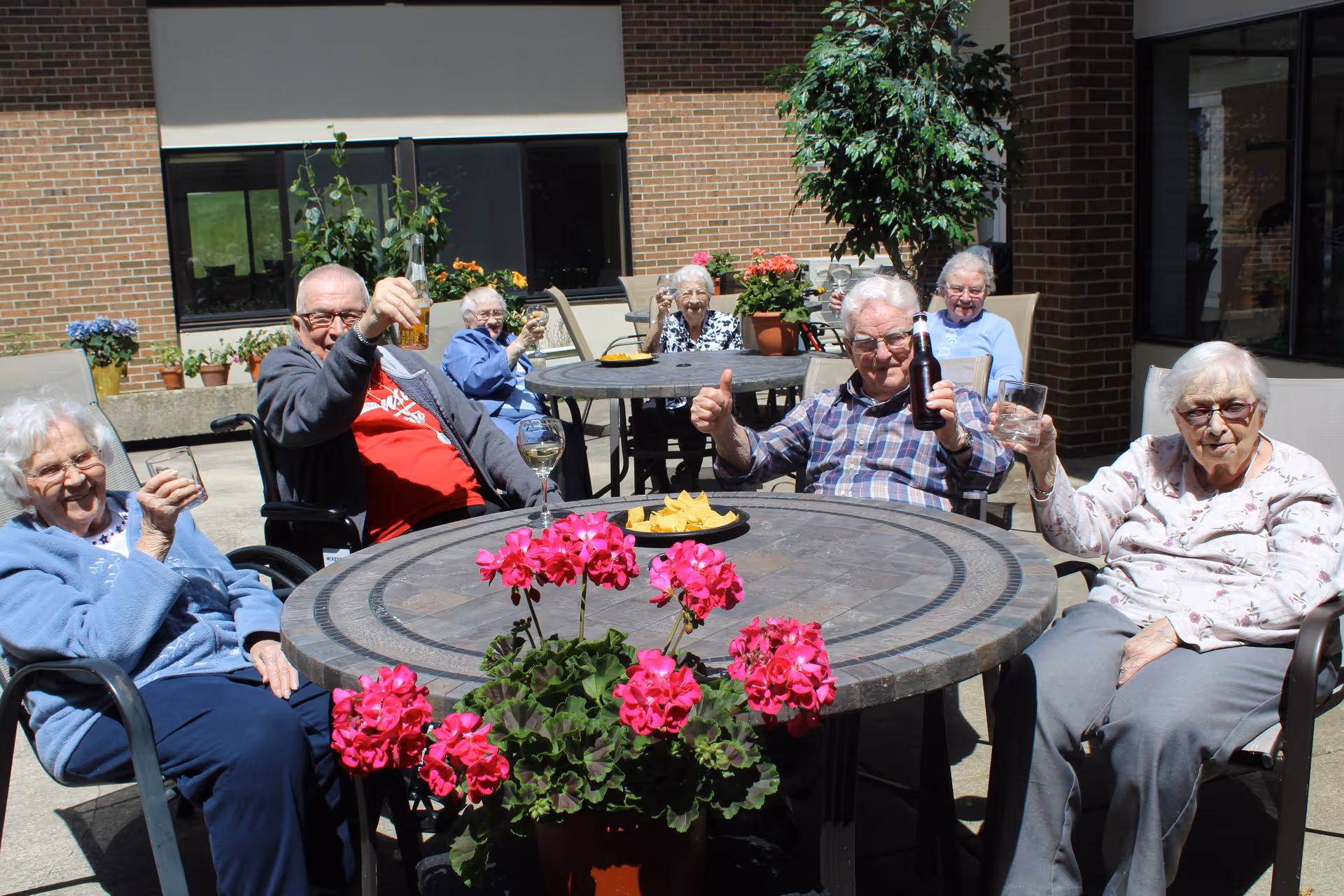 A group of elderly people sitting outdoors around two circular tables with pink flowers in the foreground. They are smiling, holding drinks, and enjoying a sunny day at a senior living facility with brick walls and windows in the background.