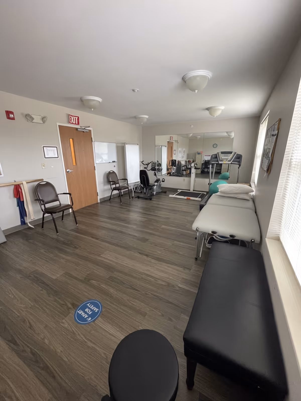 A small exercise room with wooden flooring, two chairs, a stationary bike, a treadmill, a padded treatment table with pillows, a black bench, and a large mirror on the far wall. There is a door with an exit sign above it, a whiteboard, and a window with blinds letting in natural light.