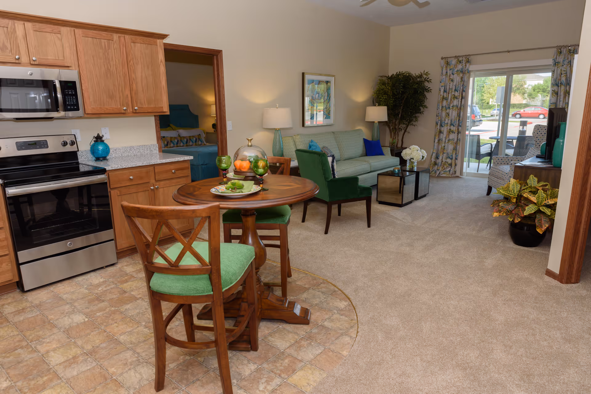 Open-plan living area with a small round dining table and green chairs, a kitchenette with stainless steel appliances, and a seating area with a sofa and sliding glass door to the outside.