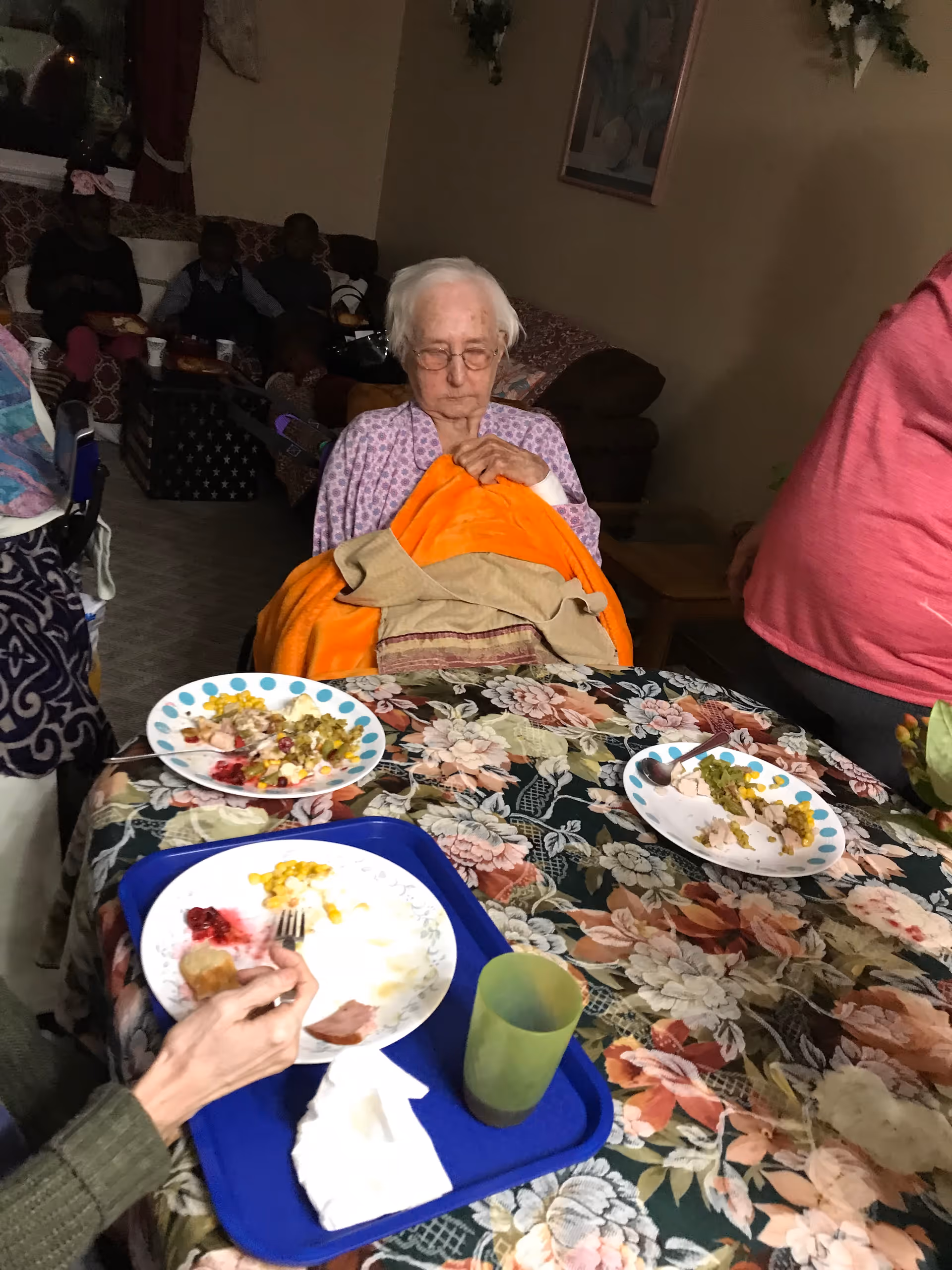 An elderly woman sitting at a table covered with a floral tablecloth, holding an orange blanket. There are plates with partially eaten food and a green cup on the table. Other people are visible in the dimly lit background, seated on couches.