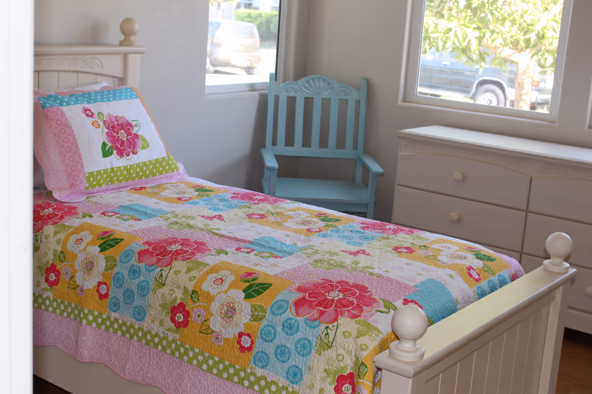 A bright bedroom with a single bed covered in a colorful floral quilt and matching pillow. Next to the bed is a light blue wooden chair, and a white dresser with drawers is positioned under two large windows letting in natural light.