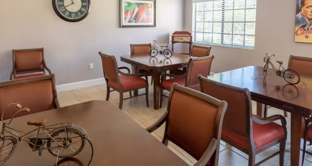 A well-lit room with several wooden tables and chairs with red cushions arranged neatly. Each table has a decorative miniature bicycle on it. There is a large window with blinds allowing natural light to enter, a clock on the wall, and framed artwork.