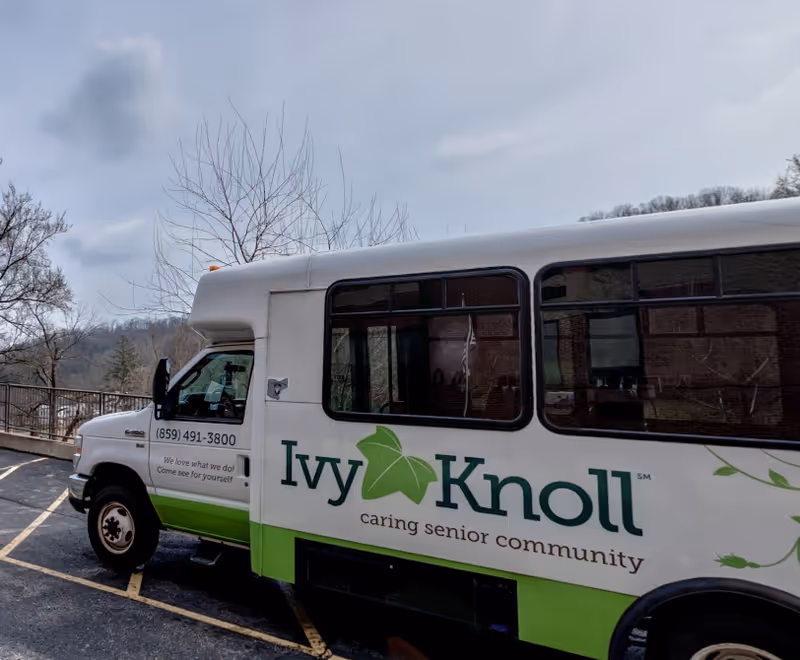 A white and green shuttle bus parked outdoors with the logo and name 'Ivy Knoll caring senior community' on its side. The bus is parked in a lot with trees and hills visible in the background under a cloudy sky.