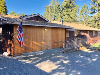 Exterior view of a single-story wooden building with an American flag mounted on the wall near the entrance. There is a wooden ramp leading to the door, surrounded by trees and clear blue sky.