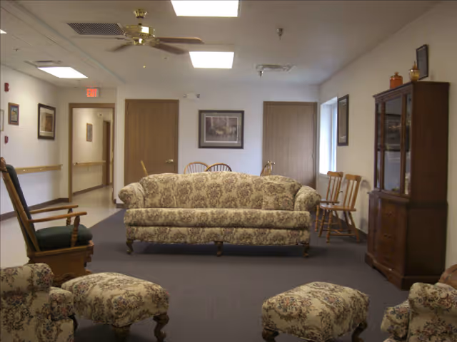 A senior living facility common area with floral patterned sofas and ottomans, wooden rocking chairs, a wooden cabinet with glass doors, and framed artwork on the walls. The room has a ceiling fan, fluorescent lighting, and a carpeted floor.