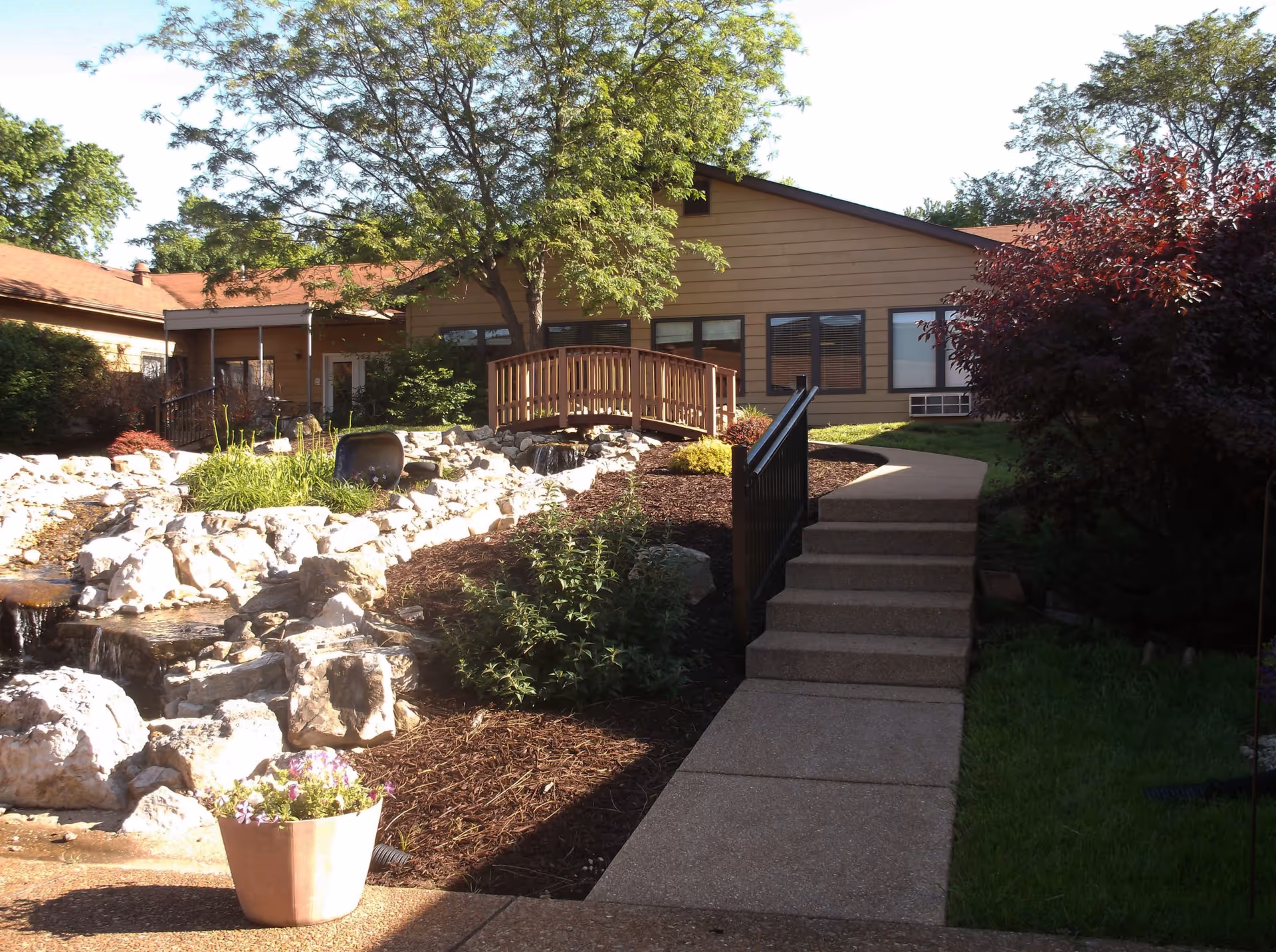 Walkway and steps leading to a small wooden bridge over landscaped rocks and a pond in front of a single-story senior living building.
