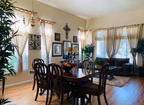 A well-lit dining room with a wooden dining table and six matching chairs. The room features large windows with sheer curtains allowing natural light to fill the space. There are decorative wall hangings, a chandelier above the table, and potted plants near the windows. A cozy sitting area with a dark sofa and a small table is visible in the background.