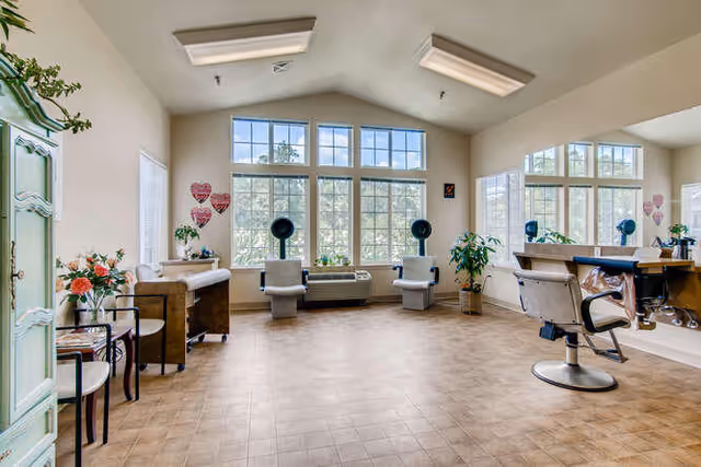 A bright and spacious hair salon room with large windows letting in natural light. The room features two salon chairs in front of a large mirror, two hair dryer chairs near the windows, a wooden cabinet with flowers, and a small table with chairs. The floor is tiled and the walls are painted light beige.